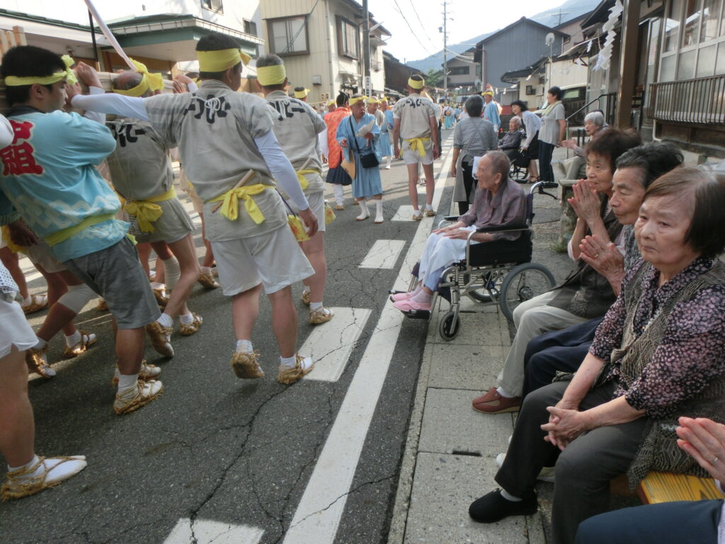 介護施設の入居者が地域のお祭りで踊りを観賞。高齢者と職員が参加する交流風景。
